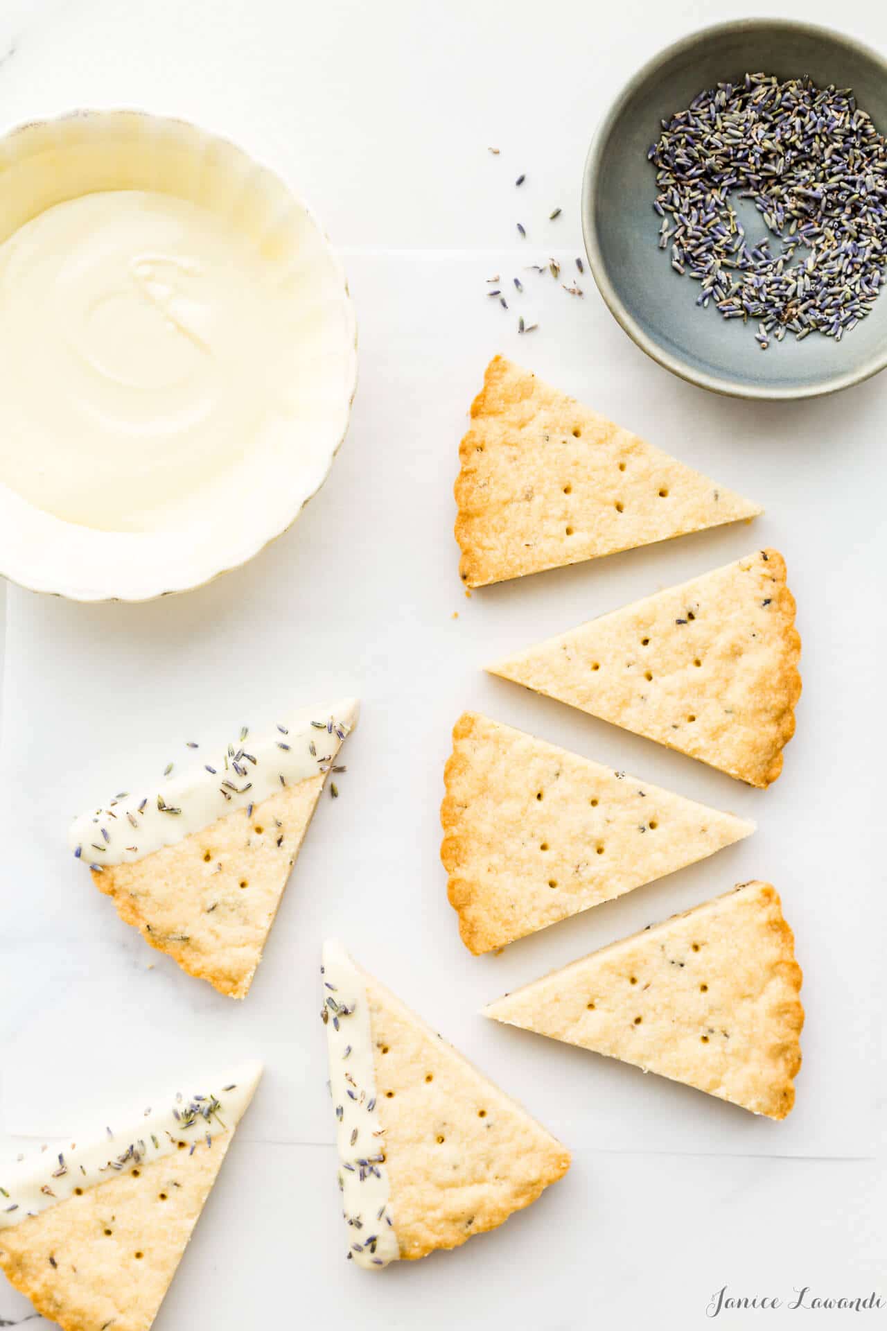 Lavender shortbread cookies dipped in a pale yellow bowl of white chocolate with a stoneware bowl of lavender