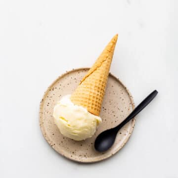 A scoop of lemon ice cream served on a speckled plate with a cone and a black spoon