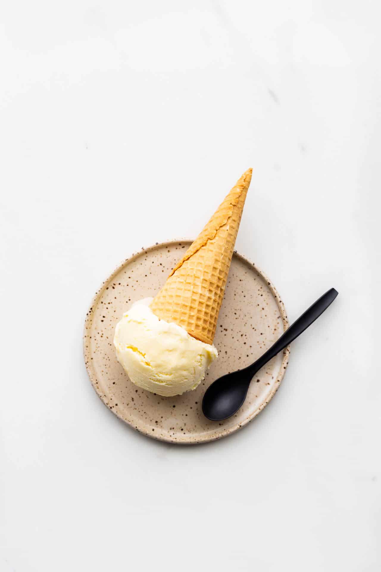 A scoop of lemon ice cream served on a speckled plate with a cone and a black spoon