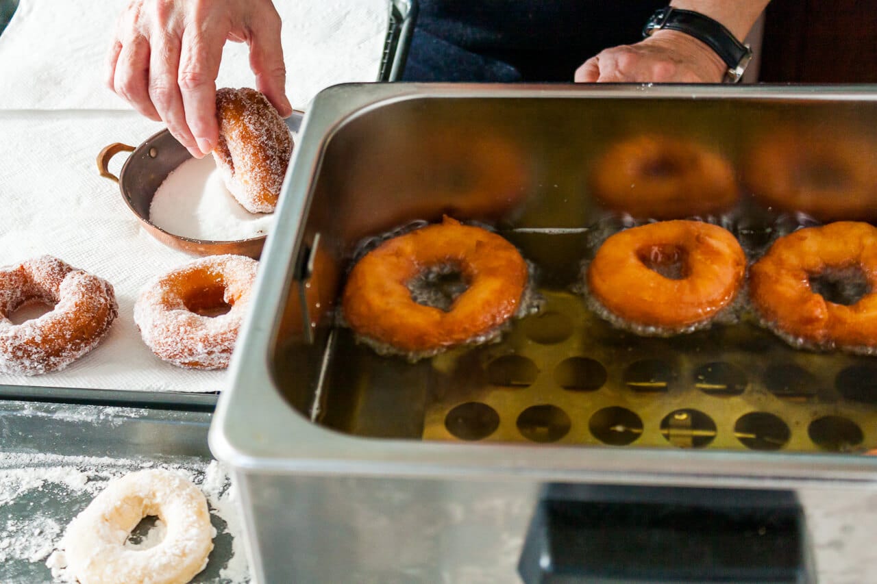 Deep frying homemade donuts in a big deep frying appliance then dipping the fried donuts in sugar to coat them before serving