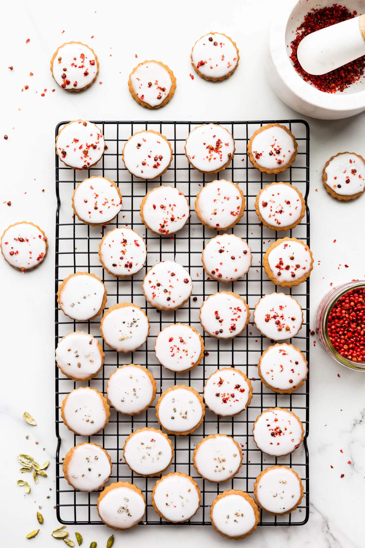 Glazed round gingerbread cookies garnished with crushed pink peppercorns and freshly ground cardamom on a black cooling rack.