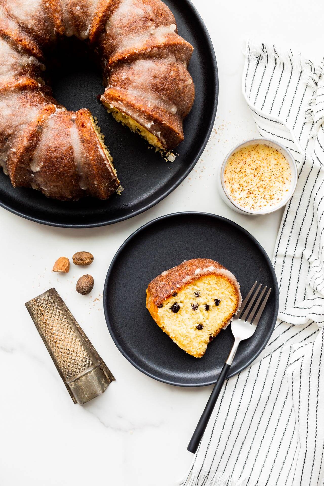 A slice of eggnog bundt cake with dried currants, served on black plate with a cup of eggnog and freshly grated nutmeg.