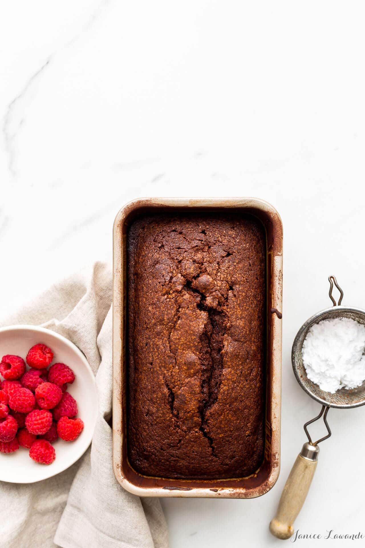Chocolate pound cake baked in a loaf cake pan, dusted with powdered sugar using a mini strainer and served with a bowl of fresh raspberries