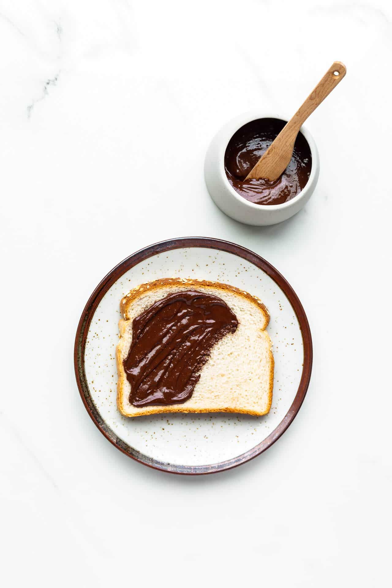 Chocolate peanut butter partly spread on a slice of white sandwich bread set on a ceramic plate with jar of spread and wood knife.