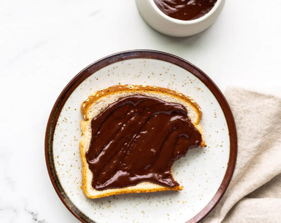 Chocolate peanut butter spread on white bread, with bite taken out. Set on a speckled ceramic plate with a beige linen napkin and a bowl of the spread with wooden knife.