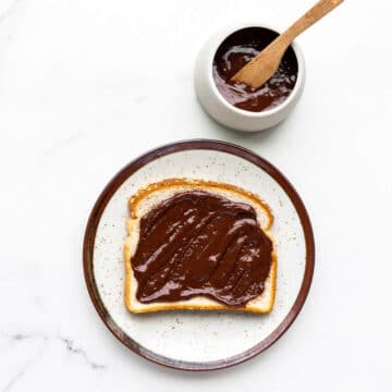 Nutella spread on white bread on a speckled ceramic plate with jar of homemade Nutella and wooden knife on side.