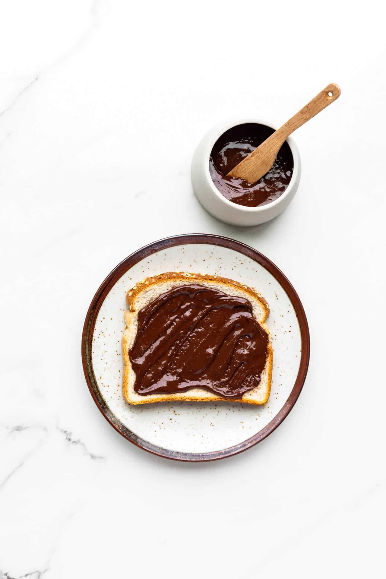 Nutella spread on white bread on a speckled ceramic plate with jar of homemade Nutella and wooden knife on side.