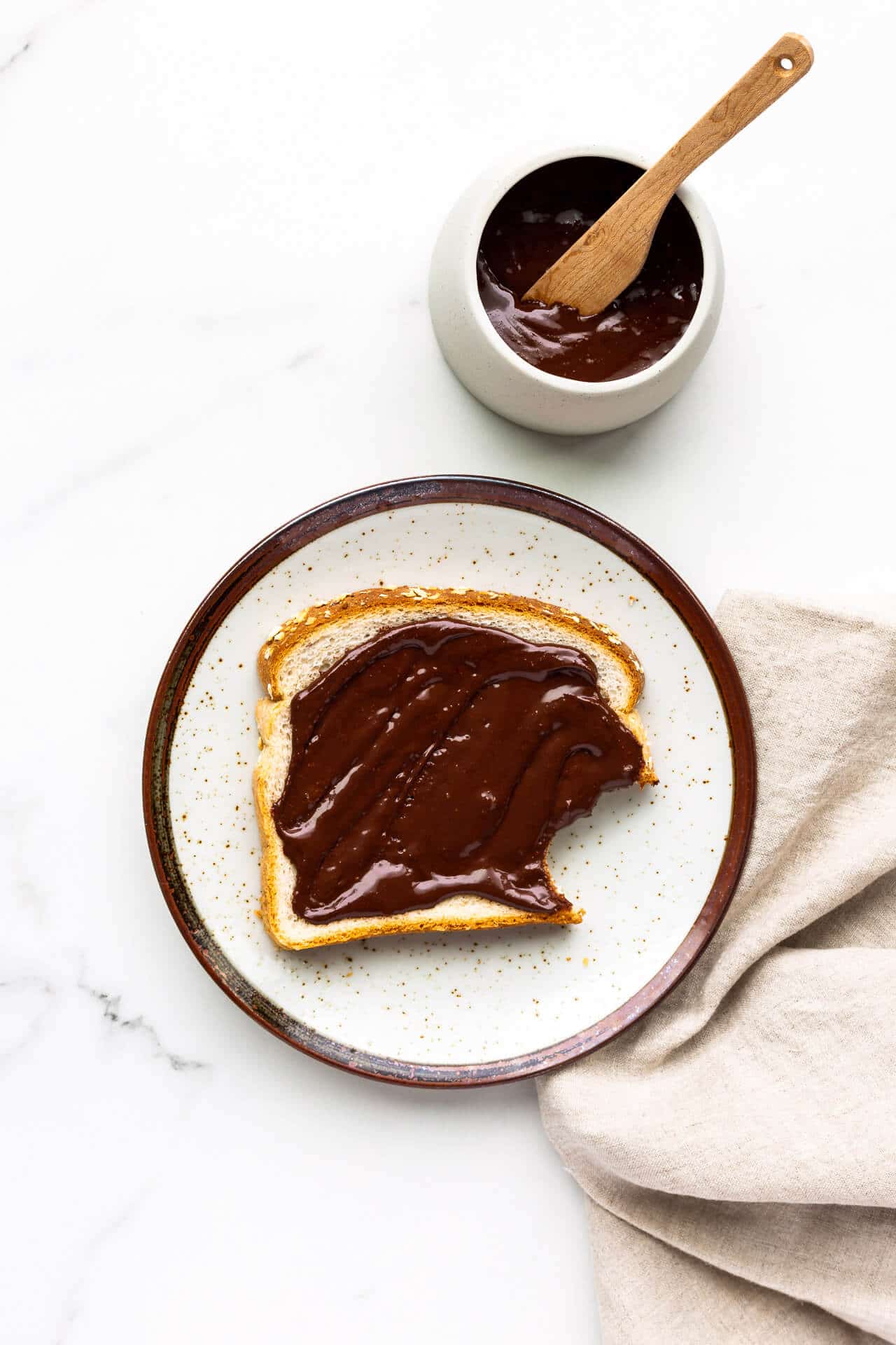 Chocolate peanut butter spread on white bread, with bite taken out. Set on a speckled ceramic plate with a beige linen napkin and a bowl of the spread with wooden knife.