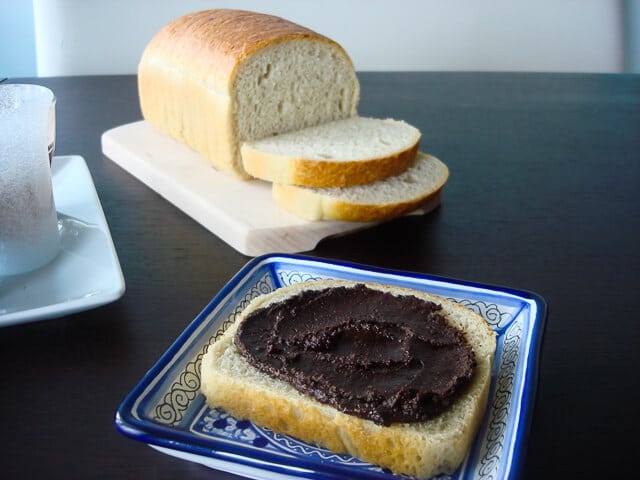 Homemade nutella smeared on a slice of white bread on a blue decorative plate with loaf in background