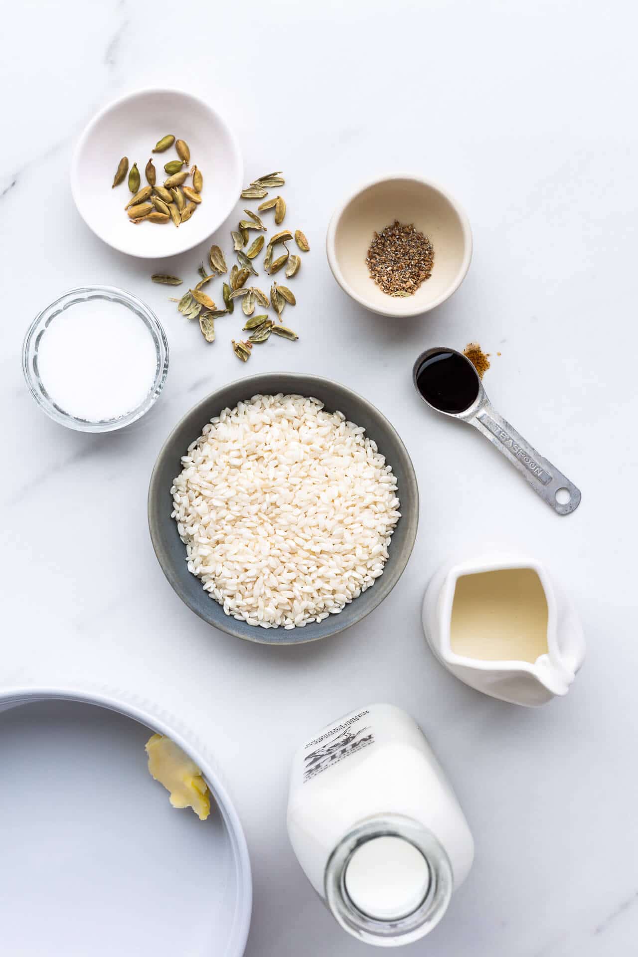 Ingredients for baked rice pudding laid out, including cardamom seeds, shelled and ground, vanilla bean paste, Arborio rice, sugar, milk, cream, and with a deep baking dish with a pat of butter on the side to grease it