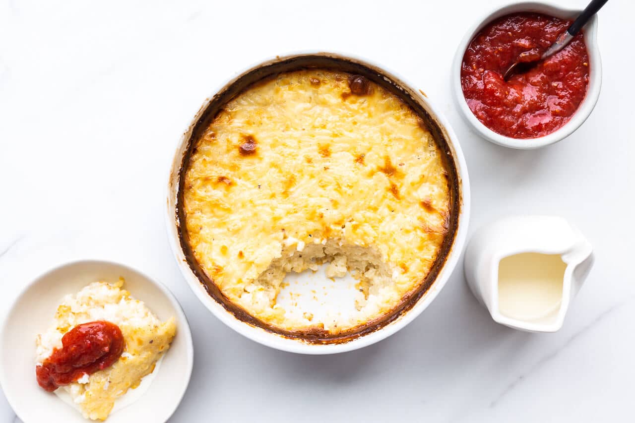 White round baking dish of baked rice pudding with golden brown edges, 1 portion missing served in a small bowl on the side, topped with rhubarb compote and cream on the side