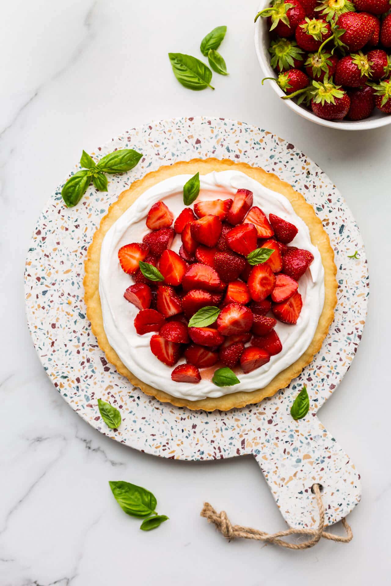 Fresh strawberry tart on a terrazzo board garnished with basil leaves and a colander of berries on the side.