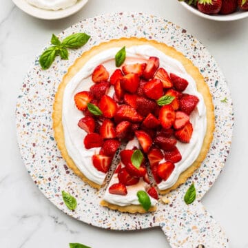 A gorgeous strawberry basil tart served on a terrazzo round board with a bowl of berries and whipped cream on the side