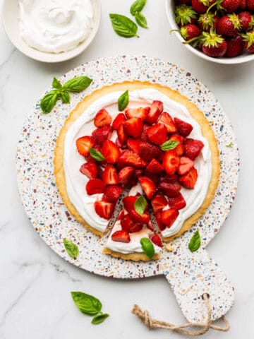 A gorgeous strawberry basil tart served on a terrazzo round board with a bowl of berries and whipped cream on the side