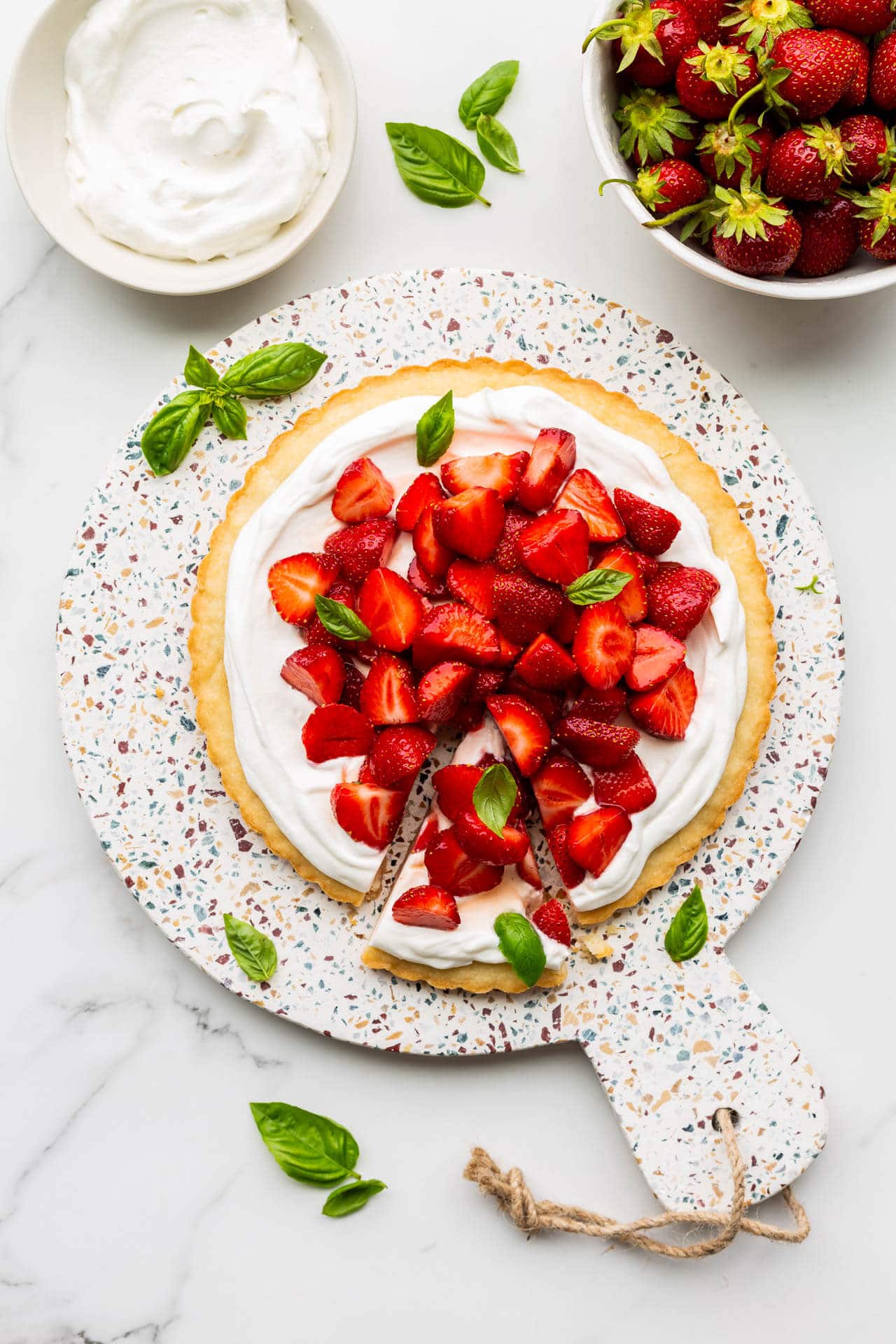 A gorgeous strawberry basil tart served on a terrazzo round board with a bowl of berries and whipped cream on the side.