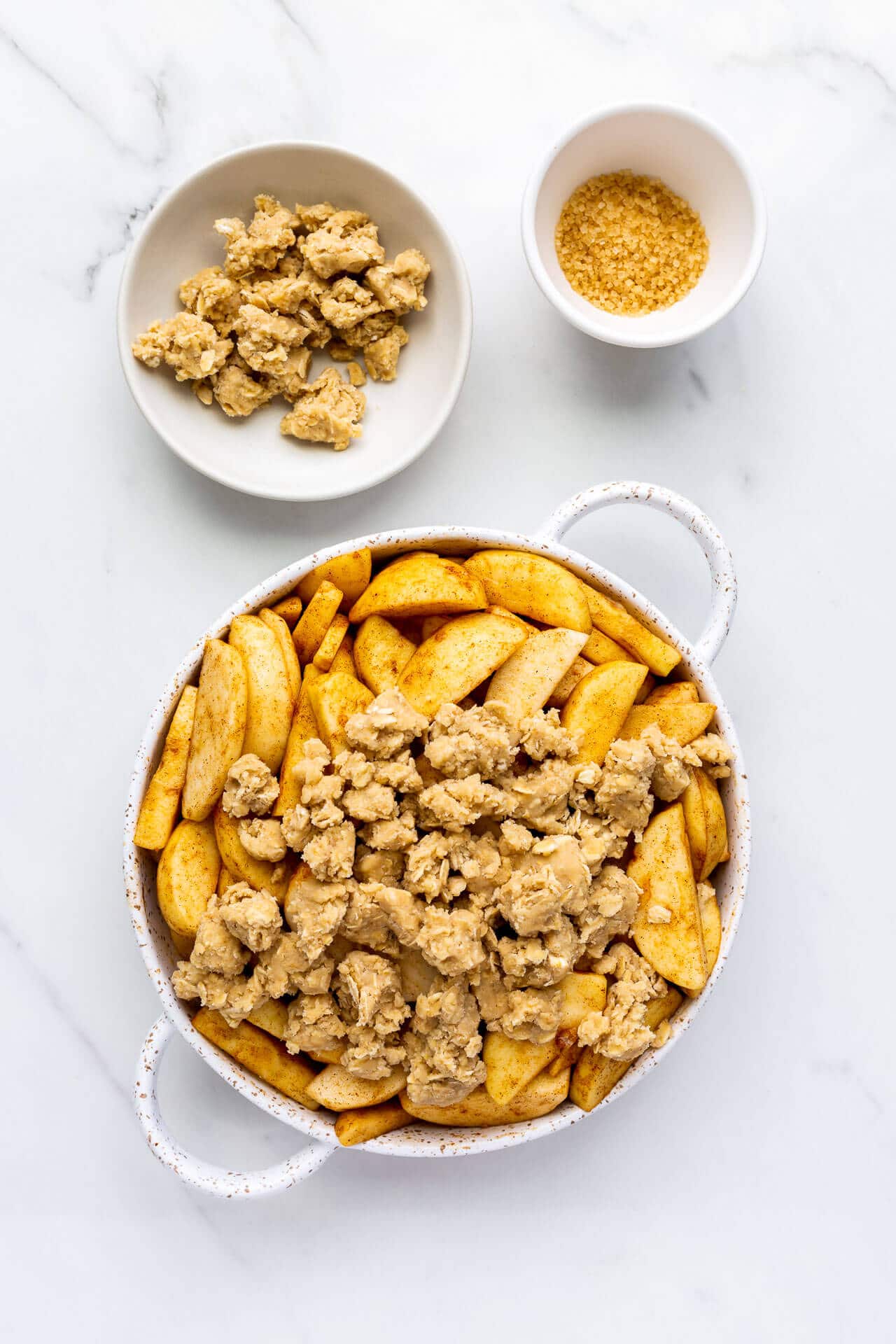 Assembling apple crisp in a white speckled bakeware dish topping with oat crumble and a little turbinado