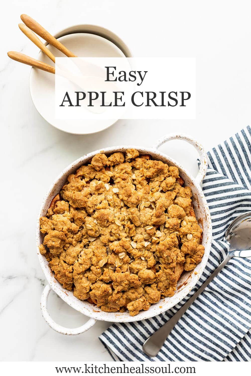 Easy apple crisp baked in a speckled white round baking dish set next to blue and white striped linen with a serving spoon, white bowls and wooden spoons