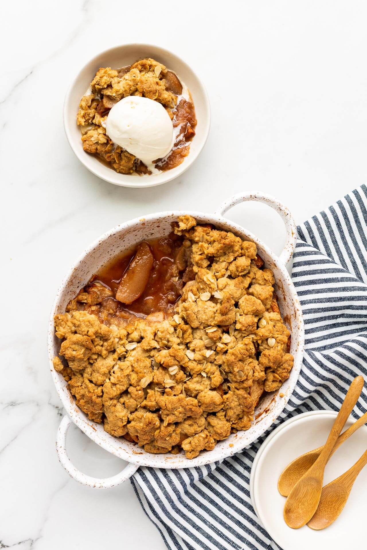 Serving a round white speckled baking dish of apple crisp into white bowls with wooden spoons and blue and white linen