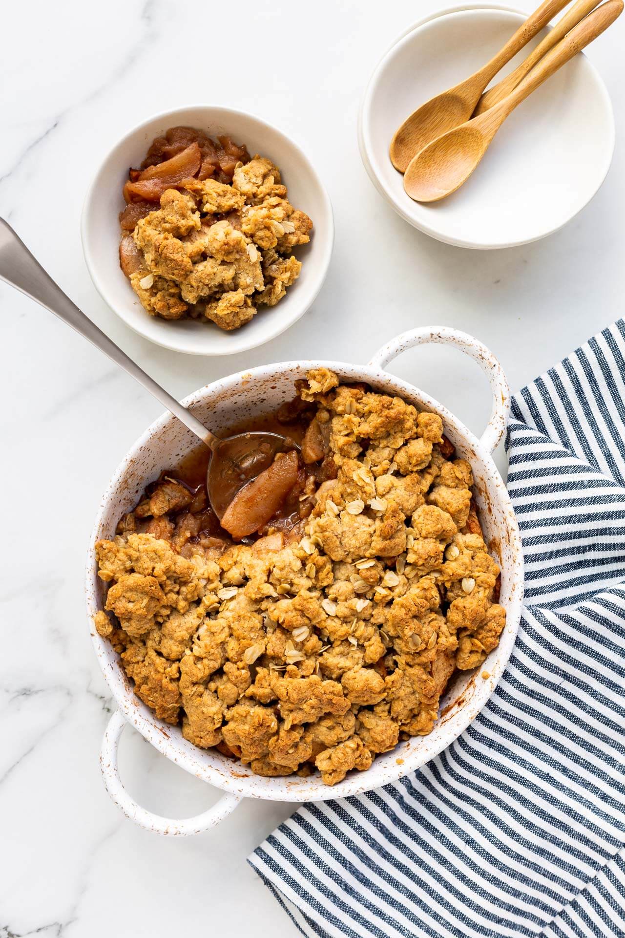 Serving a round white speckled baking dish of apple crisp into white bowls with wooden spoons and blue and white linen