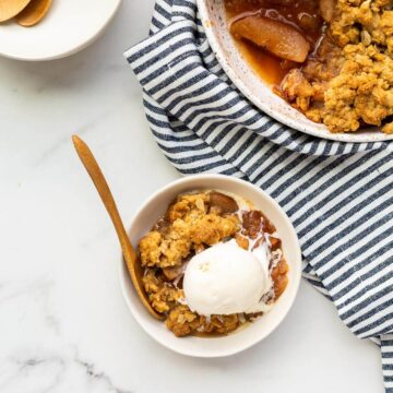 A serving of apple crisp served à la mode with a scoop of vanilla ice cream, served next to speckled dish of apple crisp, more bowls and spoons, blue and white striped linen,