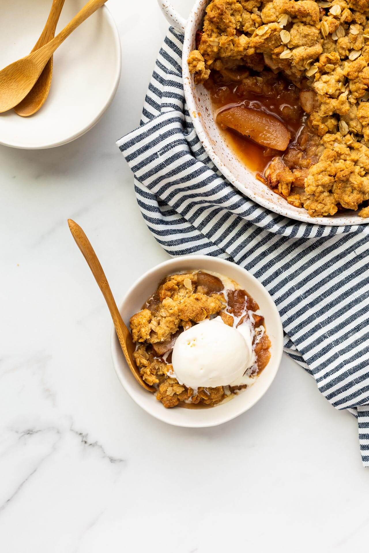 A serving of apple crisp served à la mode with a scoop of vanilla ice cream, served next to speckled dish of apple crisp, more bowls and spoons, blue and white striped linen,