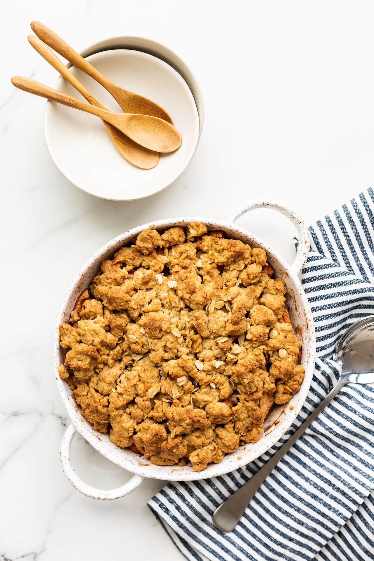 Freshly baked apple crisp in a speckled round white baking dish with handles, a blue and white striped linen, serving spoon, as well as white bowls and wooden spoons