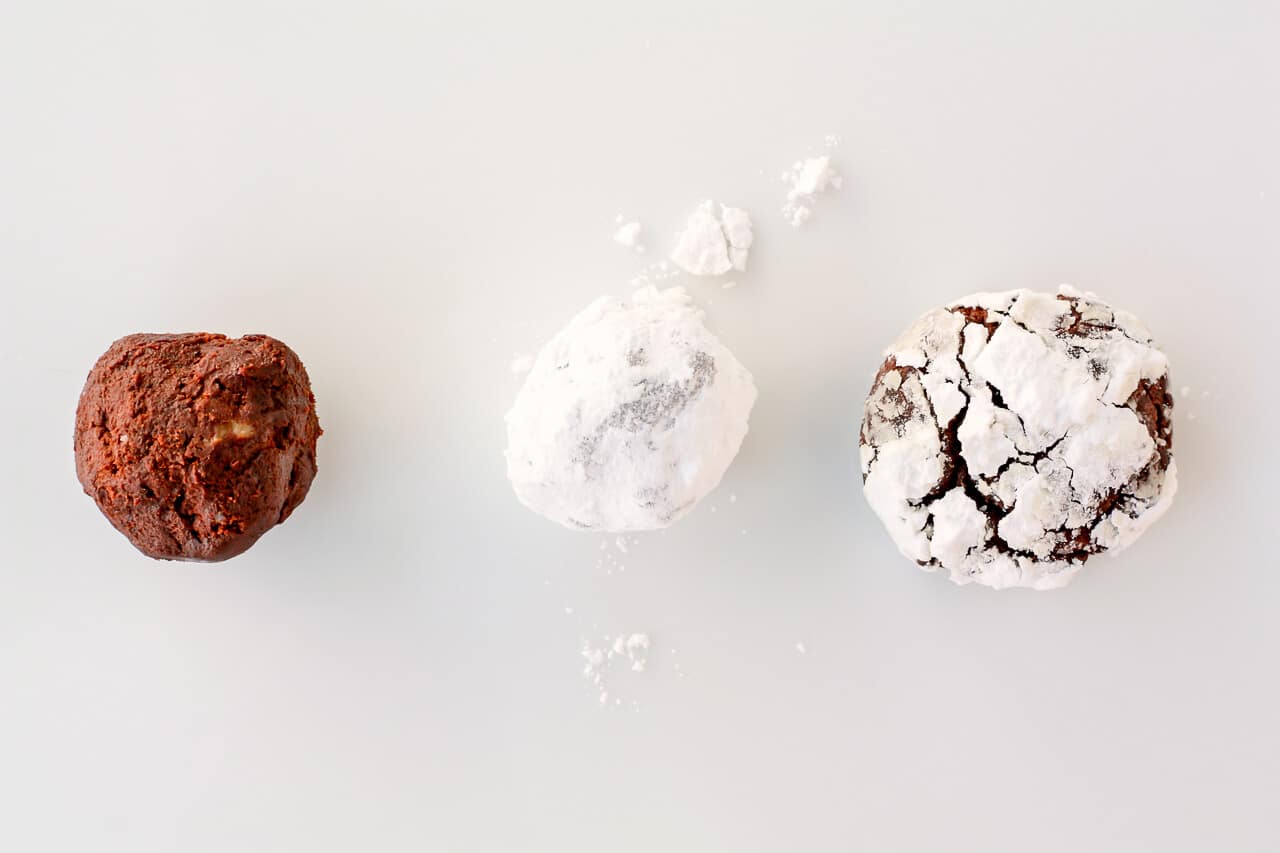 A ball of chocolate cookie dough to show what it looks raw next to a ball coated in powdered sugar before baking next to a baked chocolate crinkle cookie to show the cracked effect on the surface of the cookie.