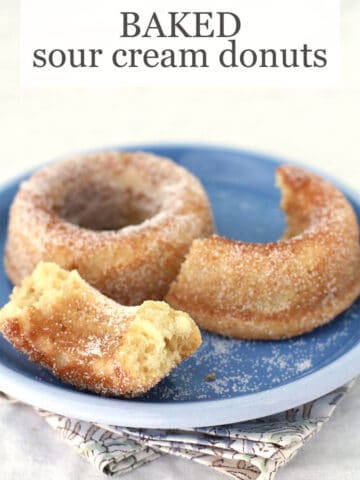 Baked sour cream donuts, one broken in half, coated with granulated sugar on a blue plate with floral napkin underneath