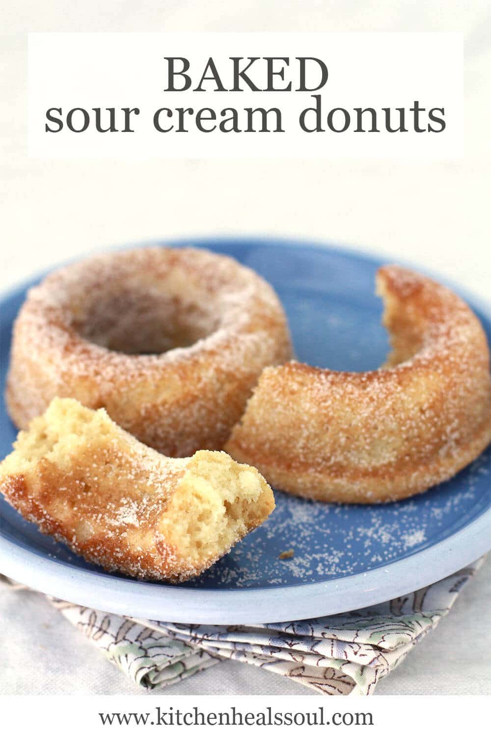 Baked sour cream donuts, one broken in half, coated with granulated sugar on a blue plate with floral napkin underneath