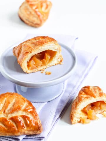 Apple turnovers with one cut open and displayed on a mini cake stand set on a purple linen napkin