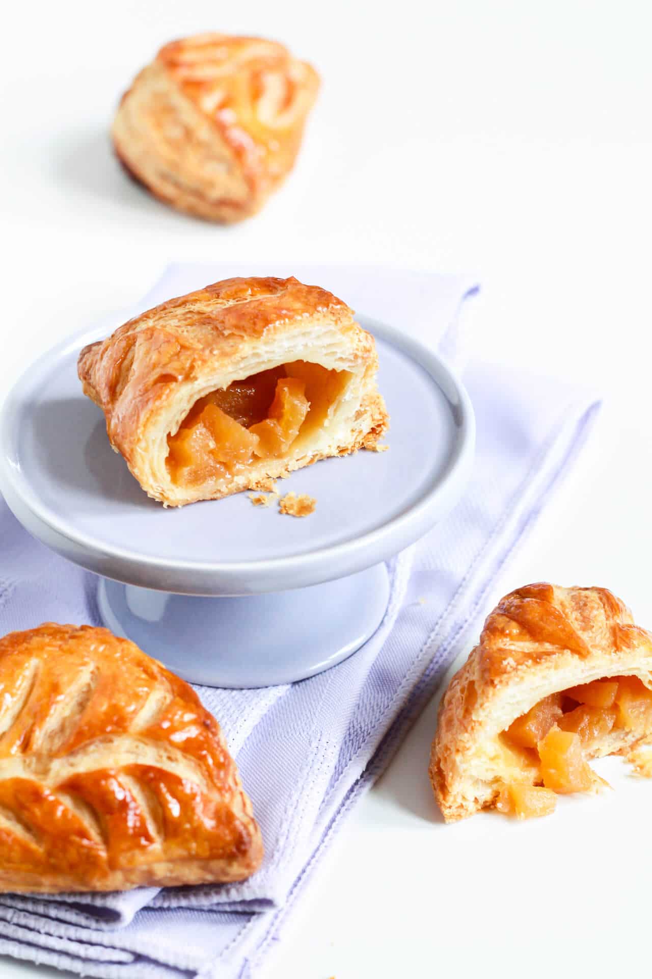 Apple turnovers with one cut open and displayed on a mini cake stand set on a purple linen napkin
