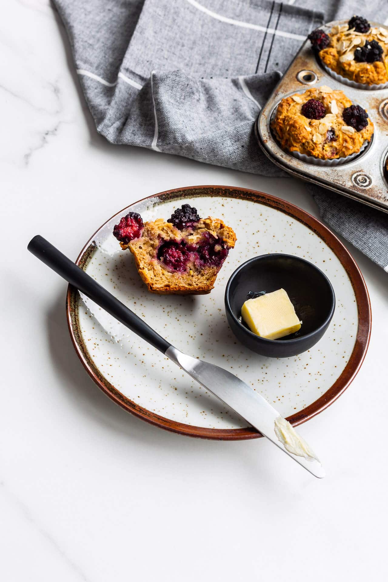 Hearty oat apple muffins with blackberries on a ceramic plate with butter and a knife, vintage muffin pan in background