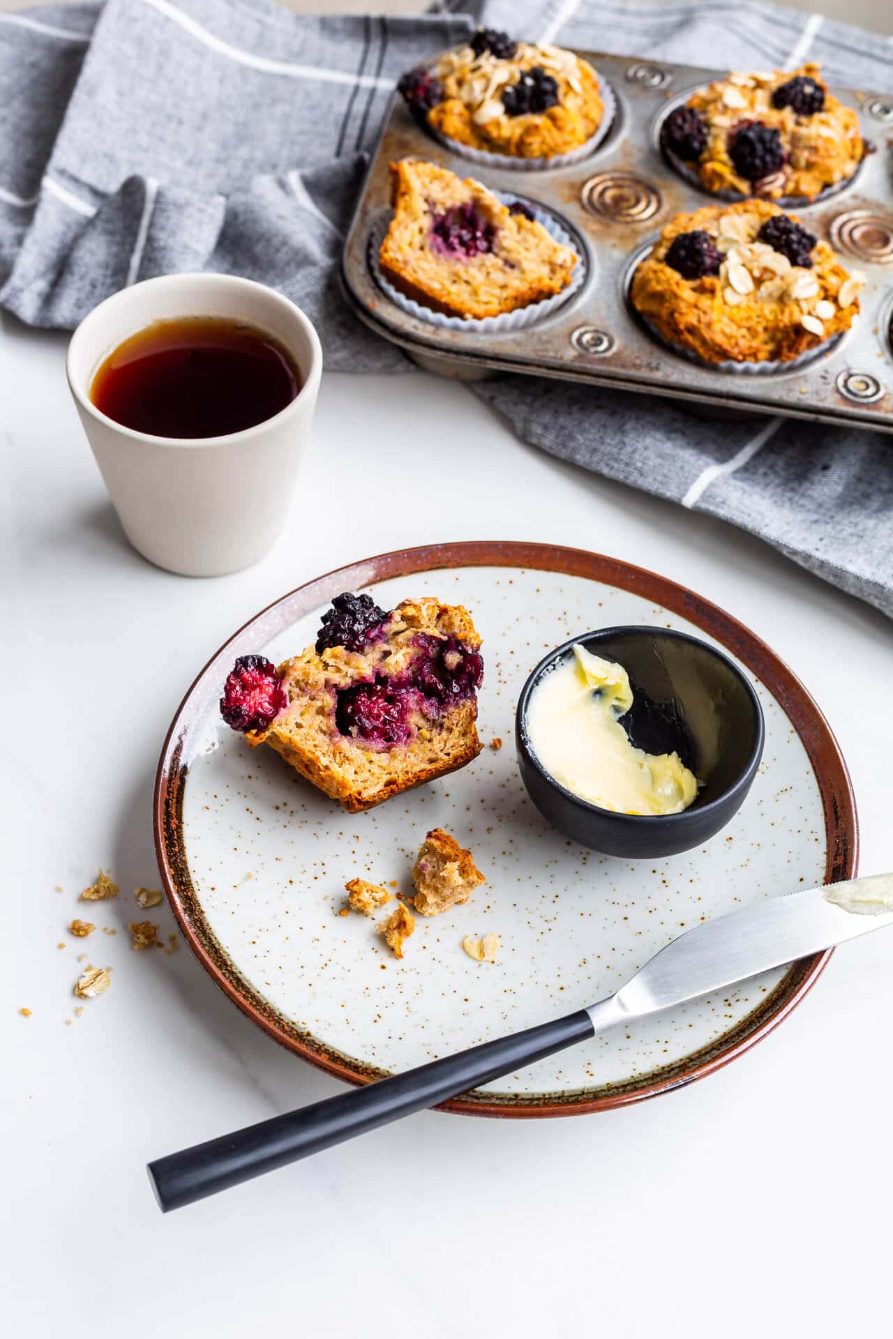 Hearty apple blackberry muffins with oats, one split open on a plate with butter and a knife, served with black tea