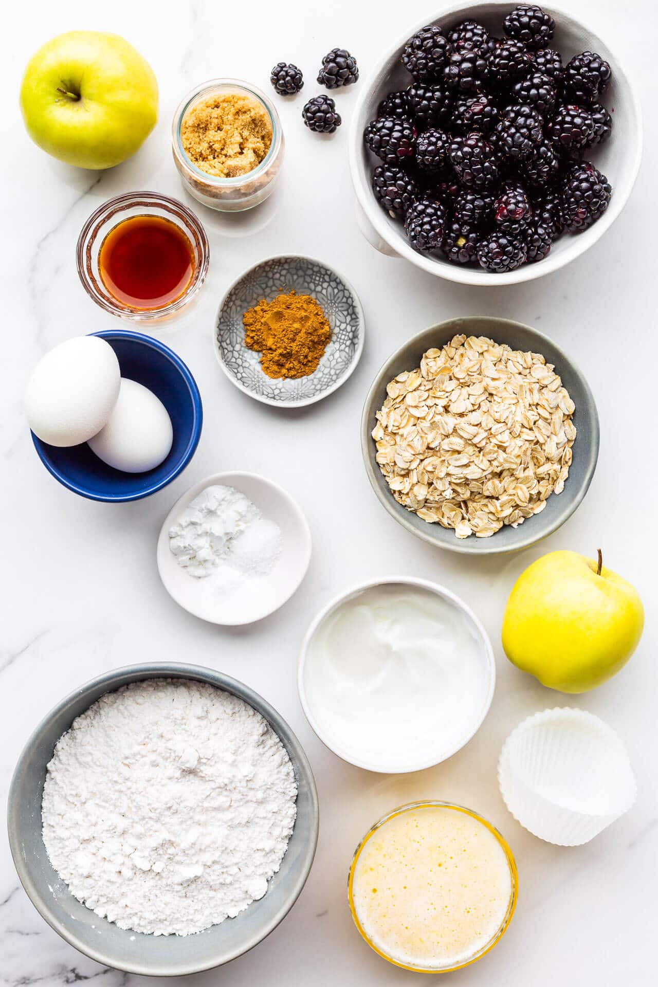 Ingredients to make apple blackberry muffins measured out and ready to be mixed, including fresh blackberries, apples, brown sugar, vanilla extract, cinnamon, 2 eggs, baking powder, baking soda, and salt, as well as oats, yogurt, melted butter, flour, and silicone liners for baking