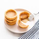 Butter tarts stacked on a beige plate with one cut open with a fork to reveal the gooey filling.