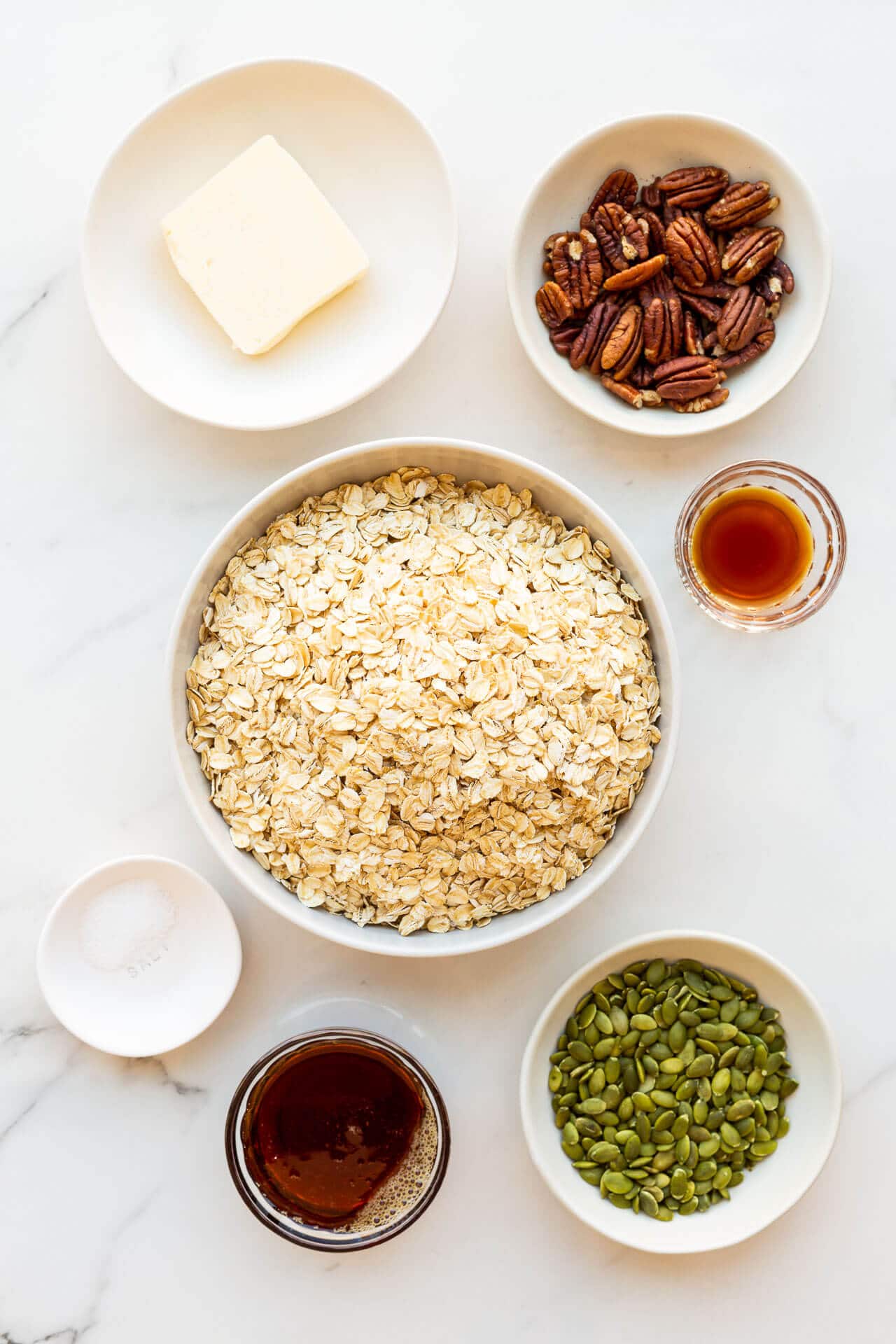 Ingredients to make granola clusters measured out in bowls and ready to be mixed: butter, pecans, vanilla extract, oats, salt, maple syrup, and pumpkin seeds