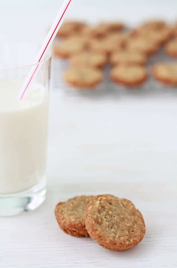 Homemade oatmeal cookies and a glass of milk with more cookies cooling on a rack in the background.