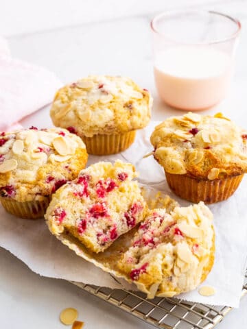 Freshly baked red currant muffins cooling on a parchment-lined wire rack with one open in half and a pink glass of milk in the back