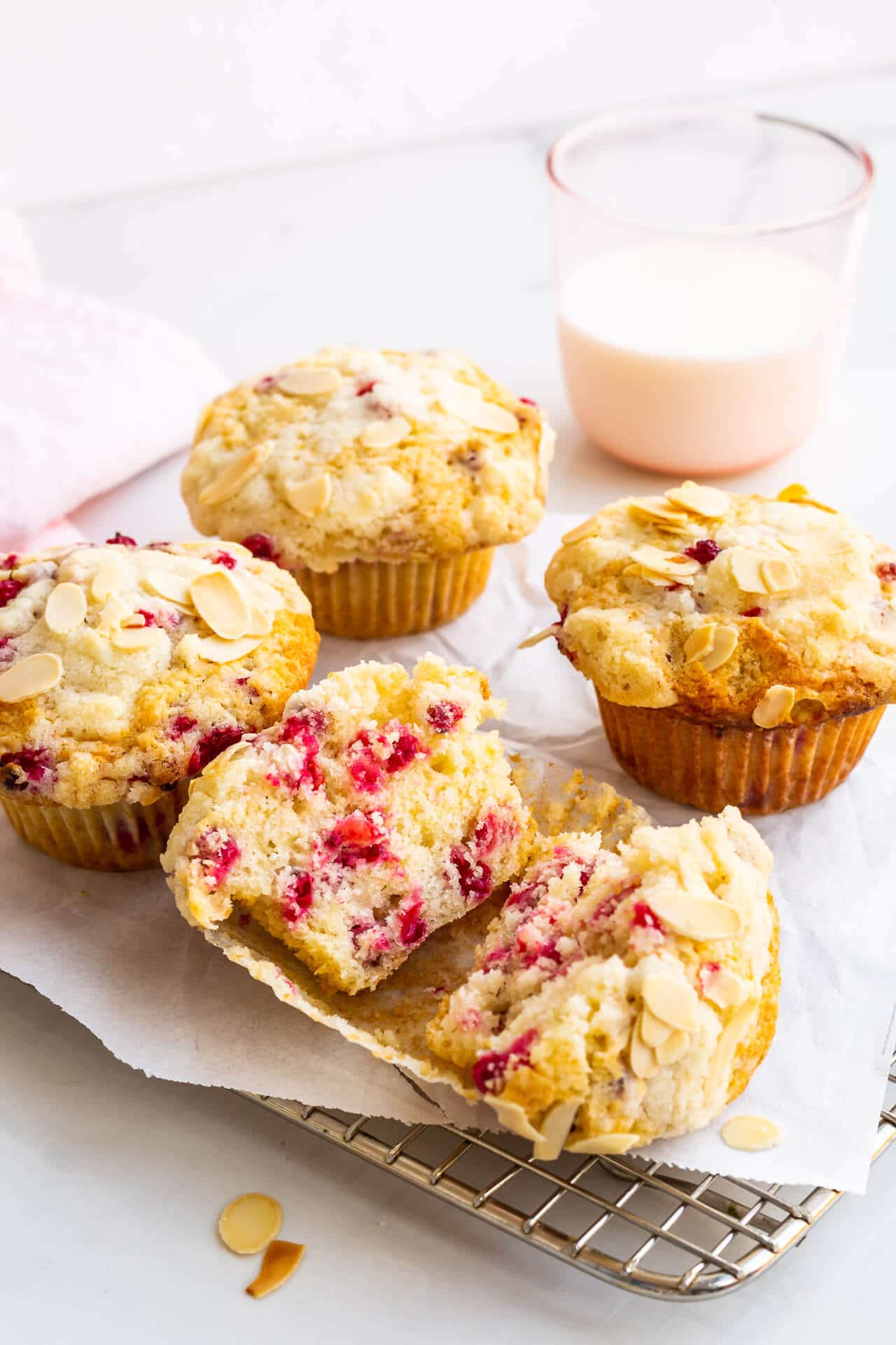 Freshly baked red currant muffins cooling on a parchment-lined wire rack with one open in half and a pink glass of milk in the back