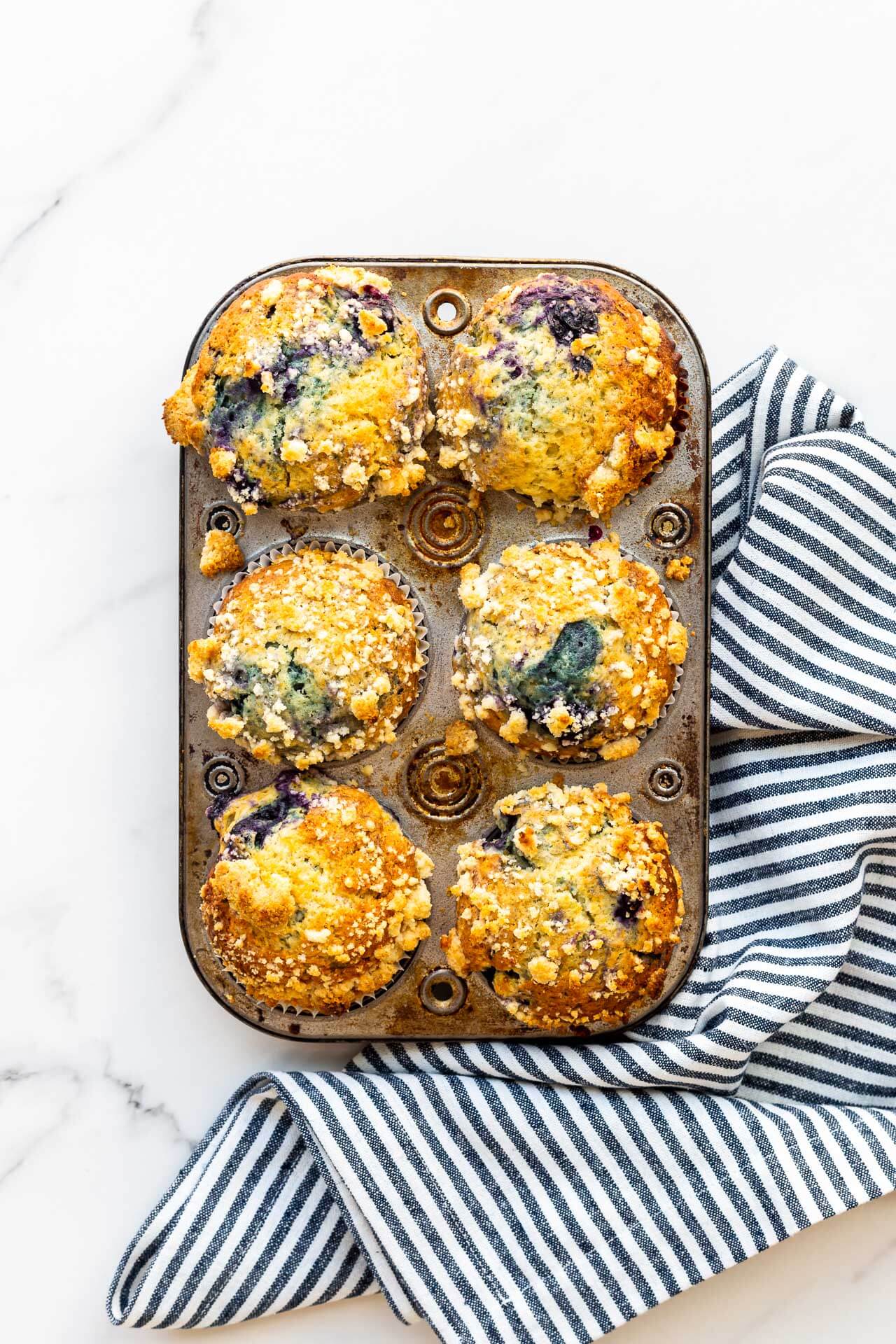 A vintage muffin pan with freshly baked blueberry streusel muffins with a stripped blue and white kitchen linen