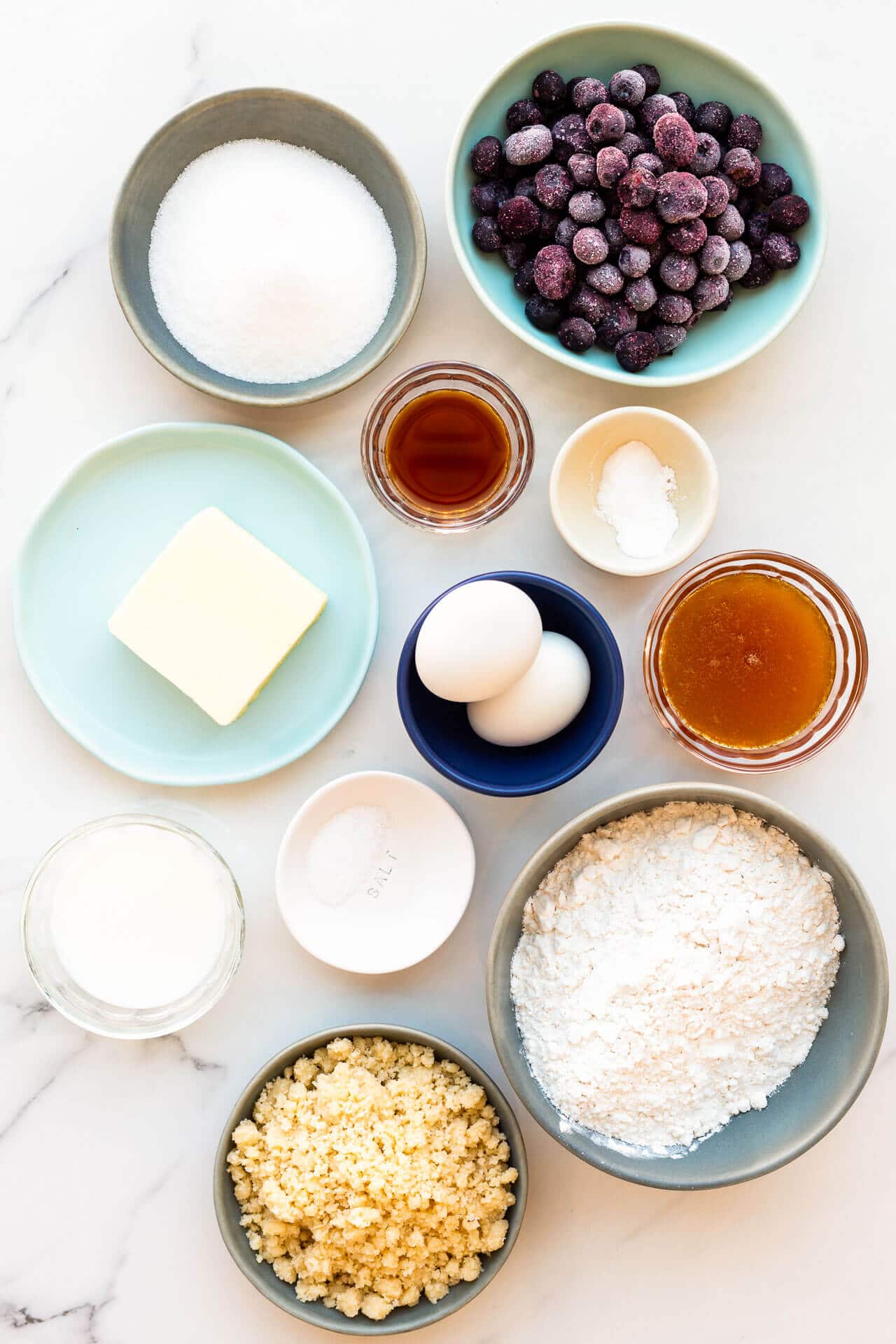 Ingredients laid out for baking blueberry muffins: sugar, frozen blueberries, vanilla, salt, honey eggs, butter, baking powder and soda, flour, and crumble topping