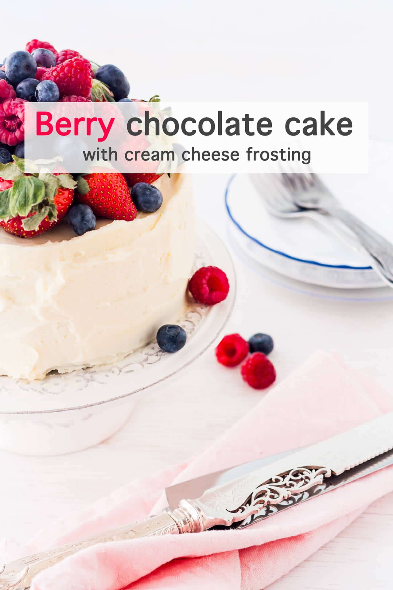 Chocolate cake with strawberries, raspberries, and blueberries, and cream cheese frosting served on a cake stand with serving knives, and a stack of plates and forks with a pink napkin