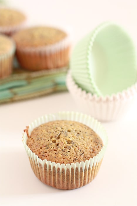 A golden brown financier in green coloured muffin paper with a stack of muffin paper liners in the background and a few more financiers