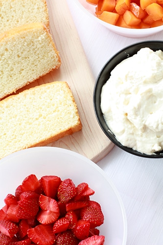 Components for assembling a trifle: sliced sponge cake, sliced strawberries, whipped cream, and chopped apricots.