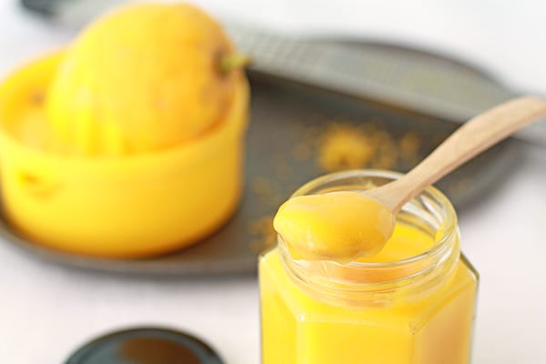 A spoonful of lemon curd sitting over a jar of it, with lemons, zester, and squeezer in the background.