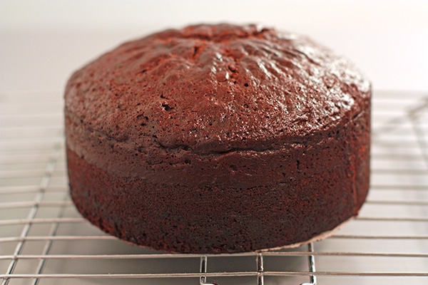 A tall chocolate cake cooling on a wire rack to show the dome that formed on top where the middle rose higher than the sides.