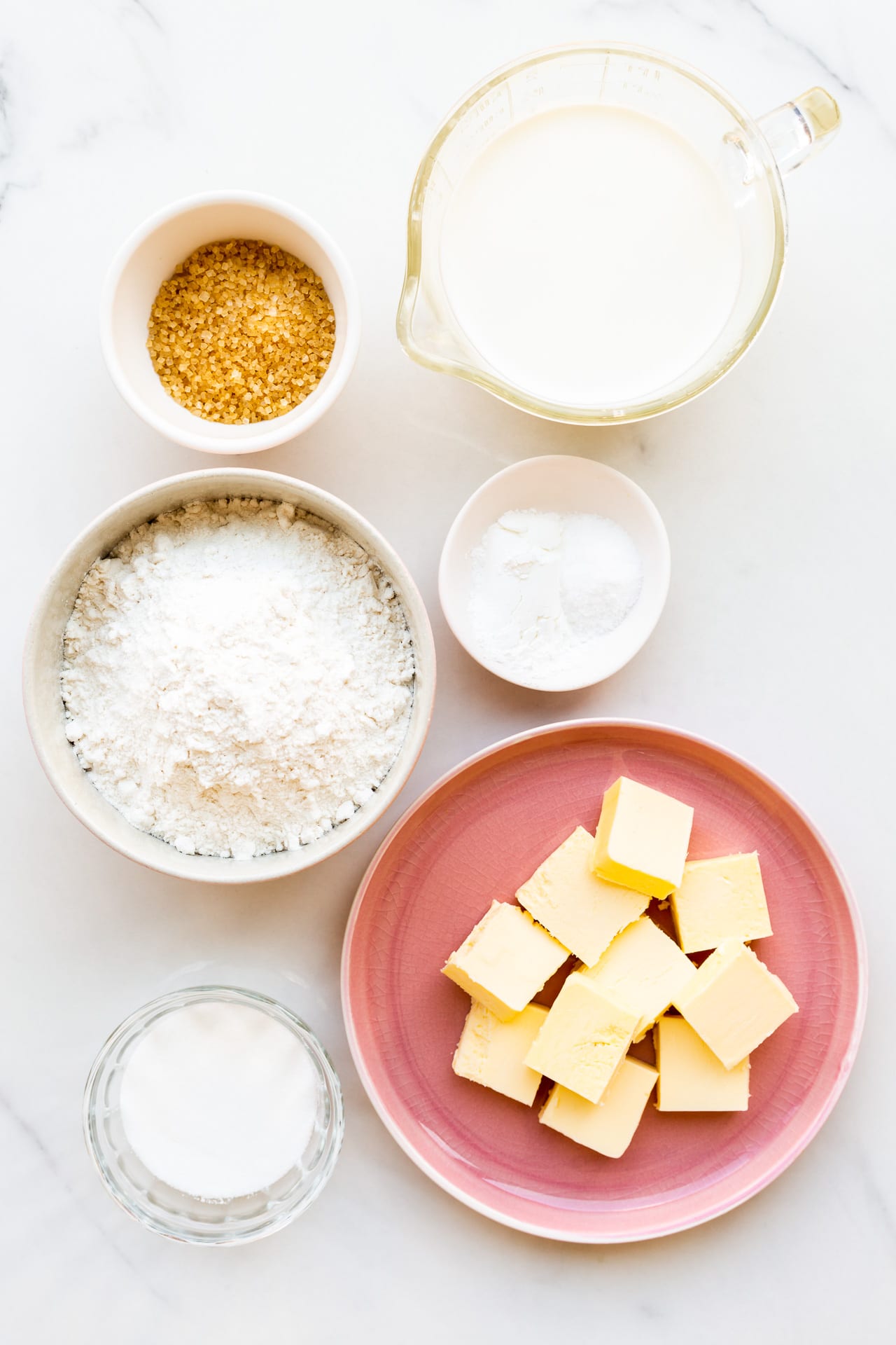 Ingredients for homemade biscuits measured out, including cubes of butter, sugar, salt, baking powder, flour, milk, and a little coarse sugar