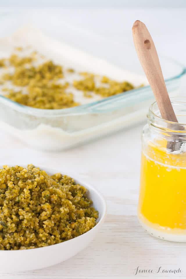 Pistachio baklava ingredients including a bowl of ground blanched pistachios so that they are extra green and melted butter with a pan of baklava in the making in the background