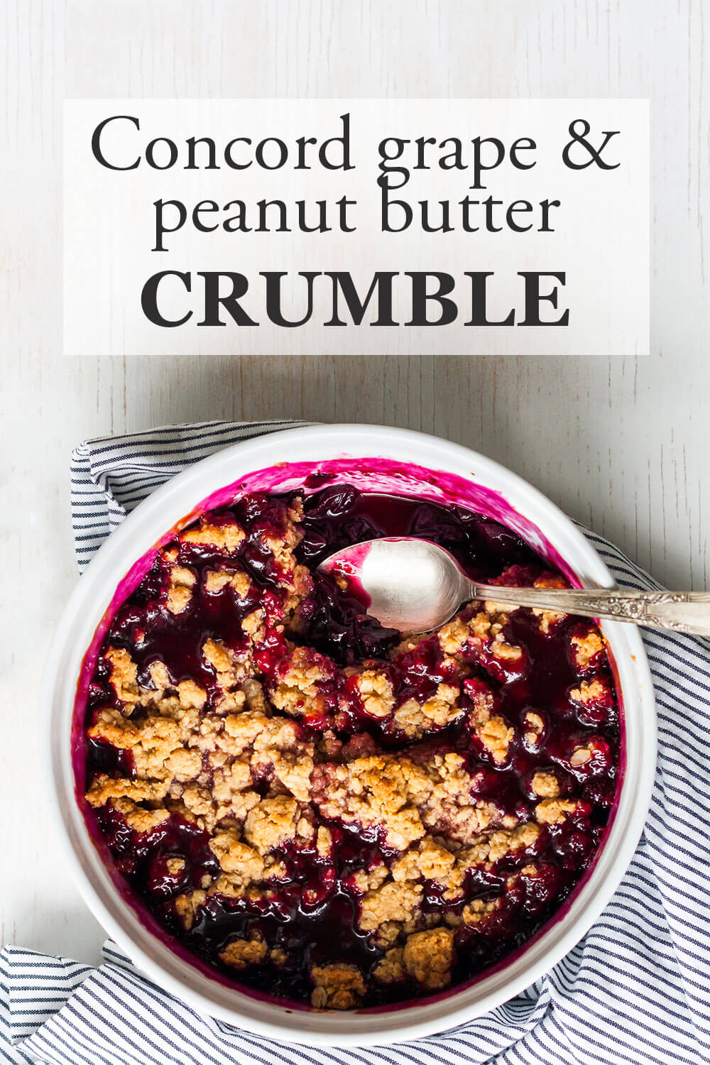 Concord grape cris with peanut butter crumble in a large white round baking dish with a silver serving spoon and a blue and white table linen