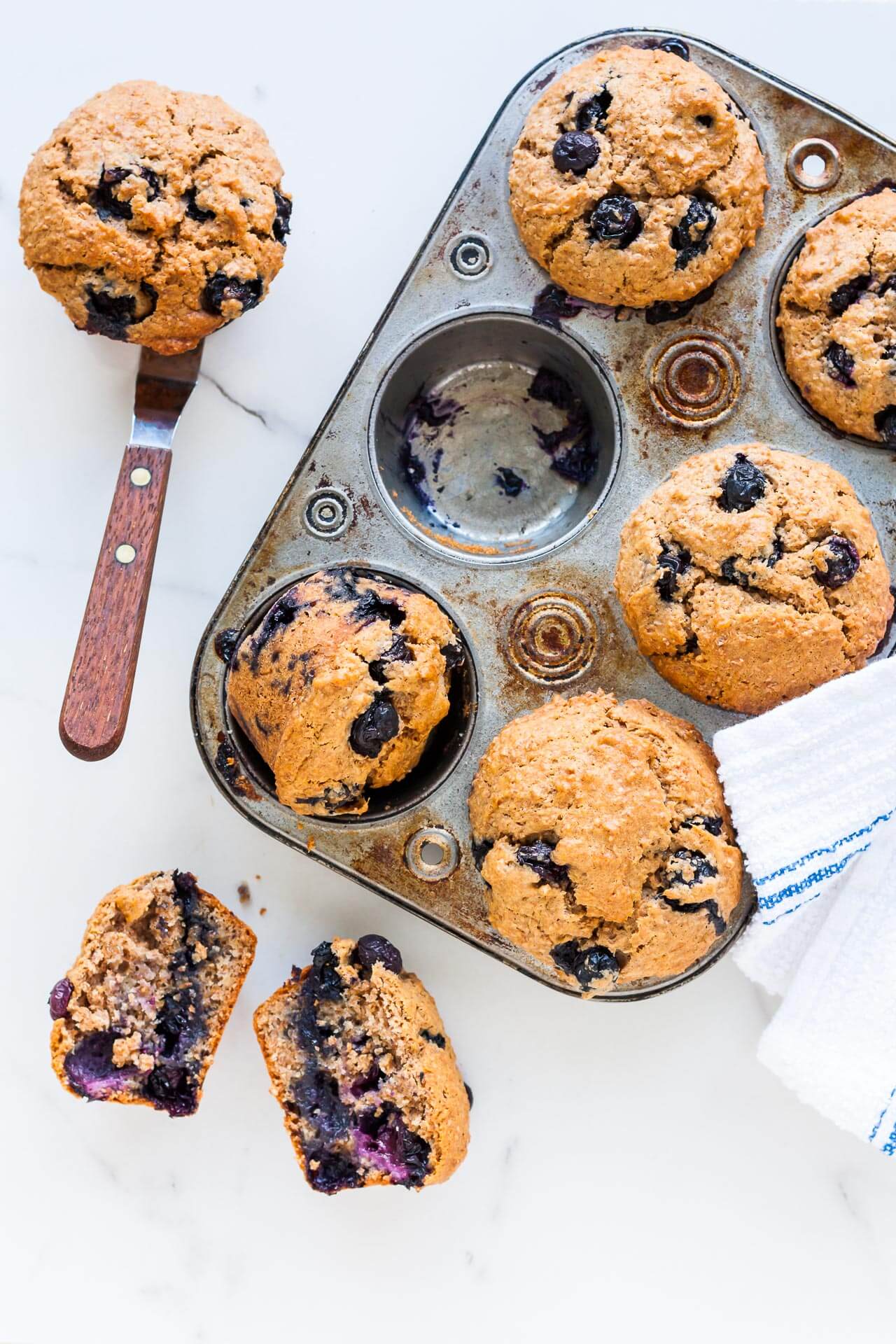 Vintage muffin pan with baked blueberry bran muffins. One cup empty and muffin is on side on an offset spatula. One muffin is broken open to show moist interior of muffin